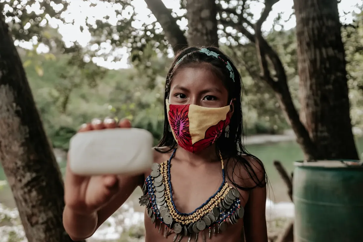 Young indigenous girl wearing a colorful face mask holds a soap bar outdoors, advocating hygiene and cultural identity.