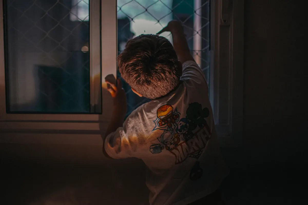 A young child gazes out a window in a dimly lit room, wearing a graphic T-shirt.