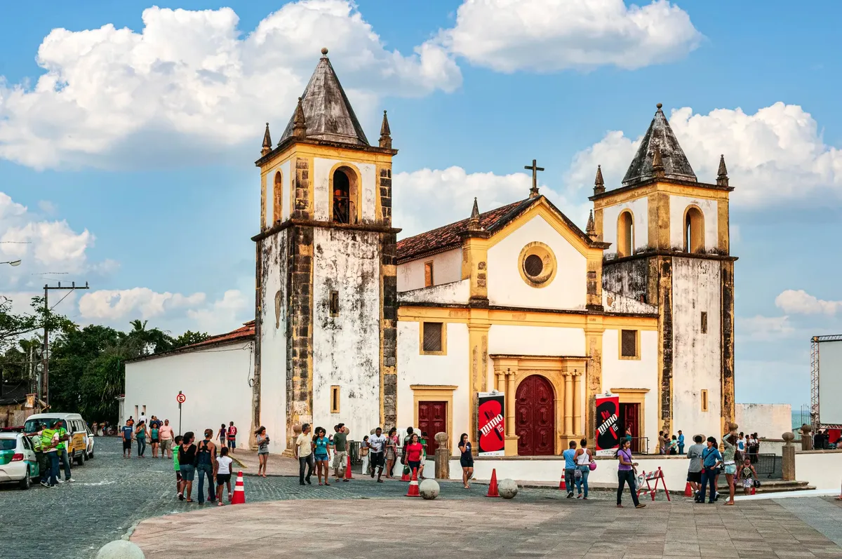 A historic church in Olinda, Brazil, with visitors gathering outside under a blue sky.