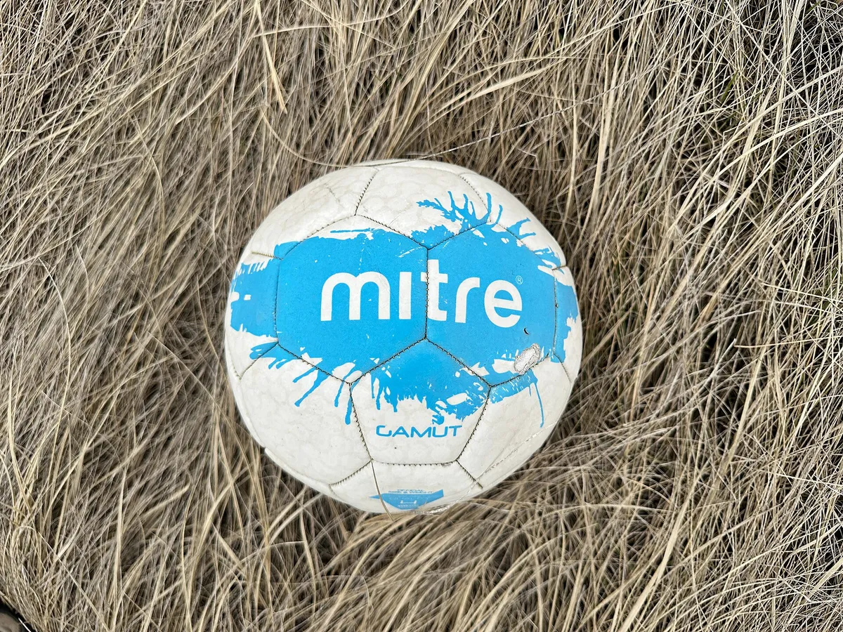 Un pallone bianco con un riquadro blu è posato sull'erba secca, visto dall'alto, senza calciatori intorno.