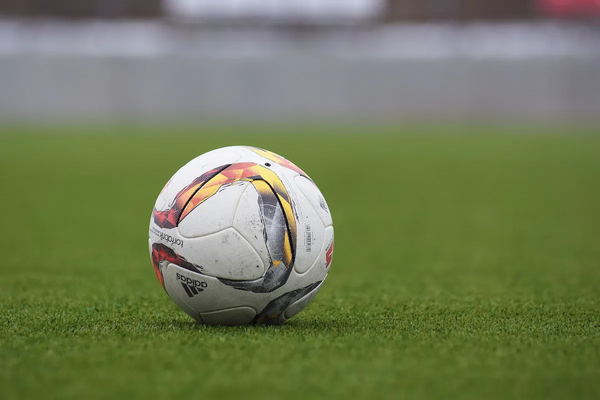 短い緑の芝の上に、黄と赤の模様が入った白いサッカーボールが一つ置かれ、後ろは灰色にぼけている。