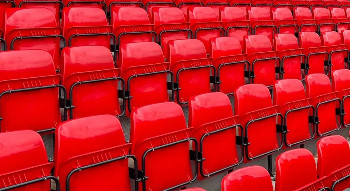 Rijen lege rode stadionstoelen met zwarte metalen beugels vullen het beeld, en er zit niemand op de tribunes.